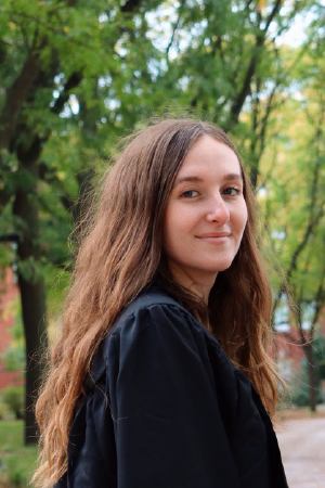 Headshot of person with long brown hair, standing outside with a black shirt.