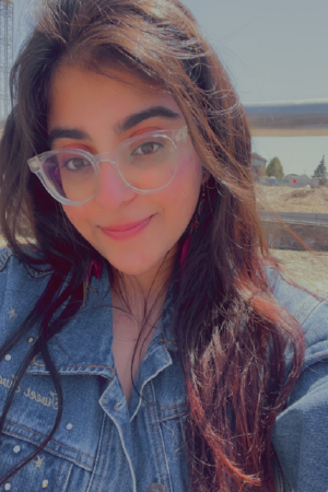 Headshot of person with long dark hair and clear-framed glasses, standing outside.