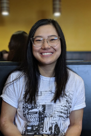 Person with long, straight black hair, wearing a white tshirt and clear framed glasses smiles. Headshot shows face, shoulders, and top half of arms and torso.