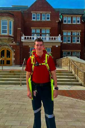 Person in a first aid outfit including a backpack, smiling, standing infront of a building outside.